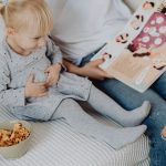 Mother reading a book to her daughter in a cozy bedroom setting with snacks nearby.